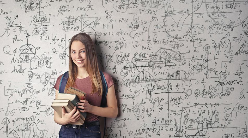 Eine junge Frau mit langen braunen Haaren steht lächelnd vor einer Wand voller mathematischer Formeln und Skizzen. Sie hält mehrere Bücher in den Armen und trägt einen Rucksack.